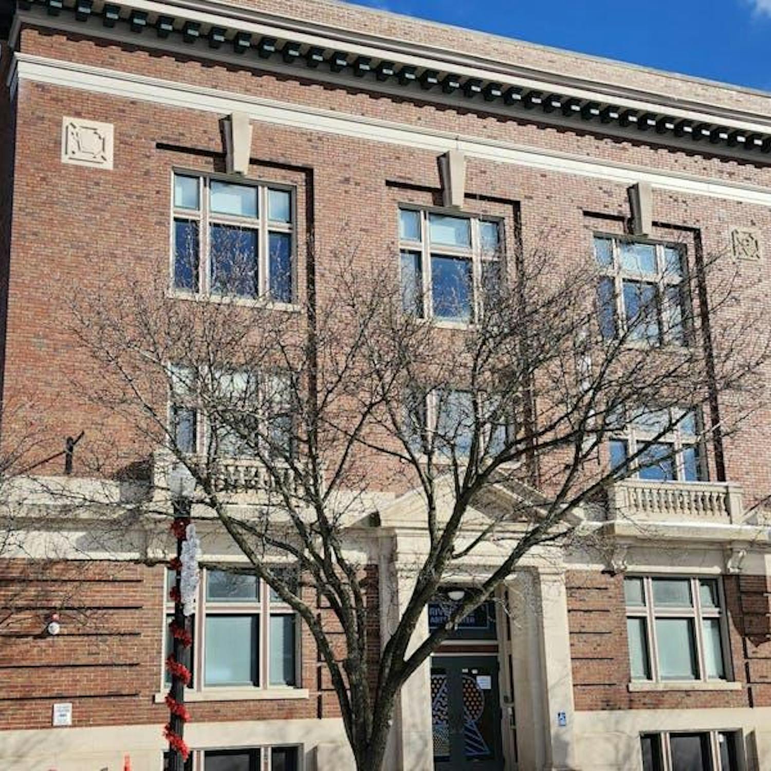 A large, rectangular-shaped brick building with several rows of windows. In front of the building is a tree that has lost all its leaves. The sky above is blue with a few white clouds, and there is another brick building to the right of the main building pictured.