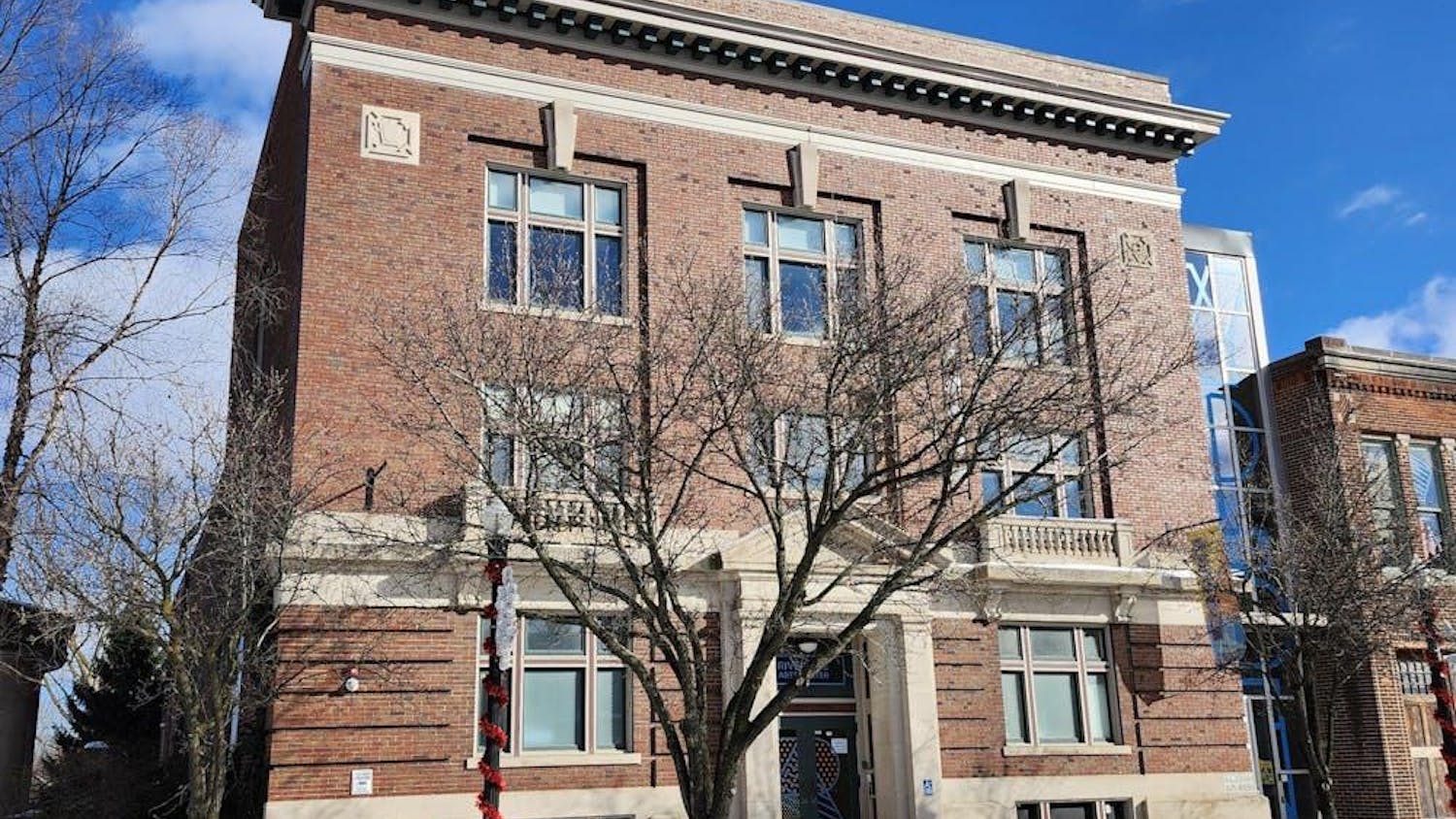 A large, rectangular-shaped brick building with several rows of windows. In front of the building is a tree that has lost all its leaves. The sky above is blue with a few white clouds, and there is another brick building to the right of the main building pictured.