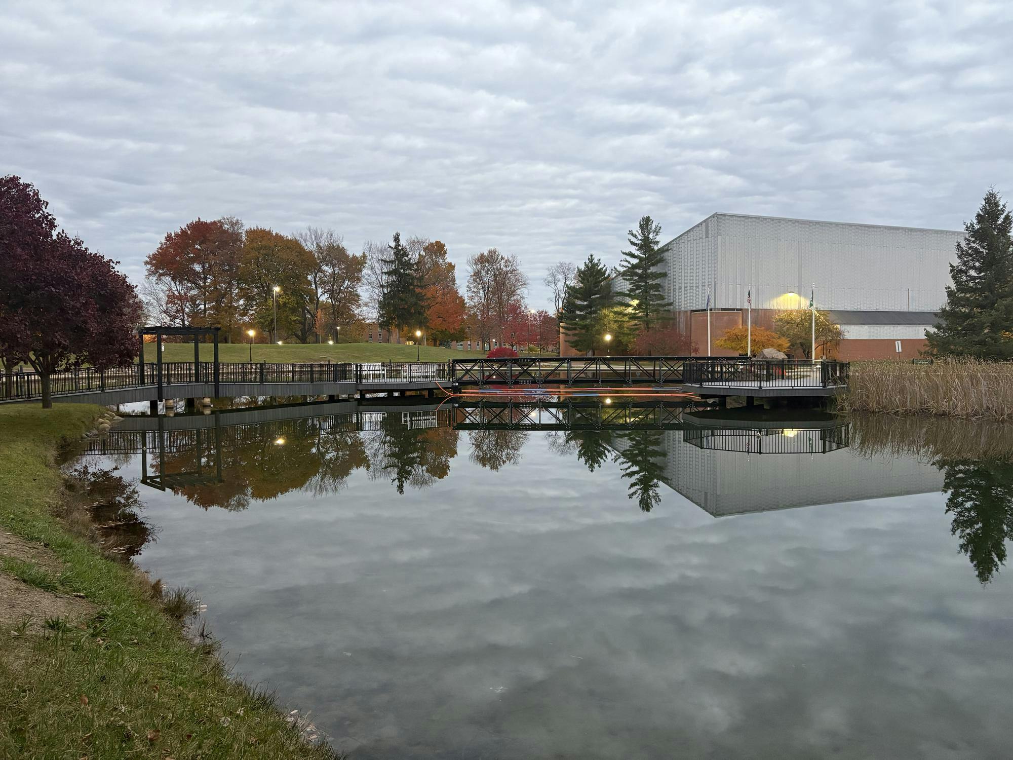 A gray and black bridge over the University Park pond with some orange cables attached to it. Black railings line the sides of the flat bridge. Overhead, the sky is overcast.