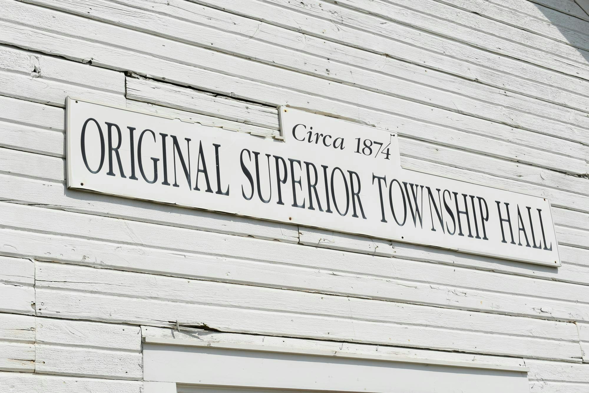 A white sign with black text indicating the original Superior Township Hall building. 