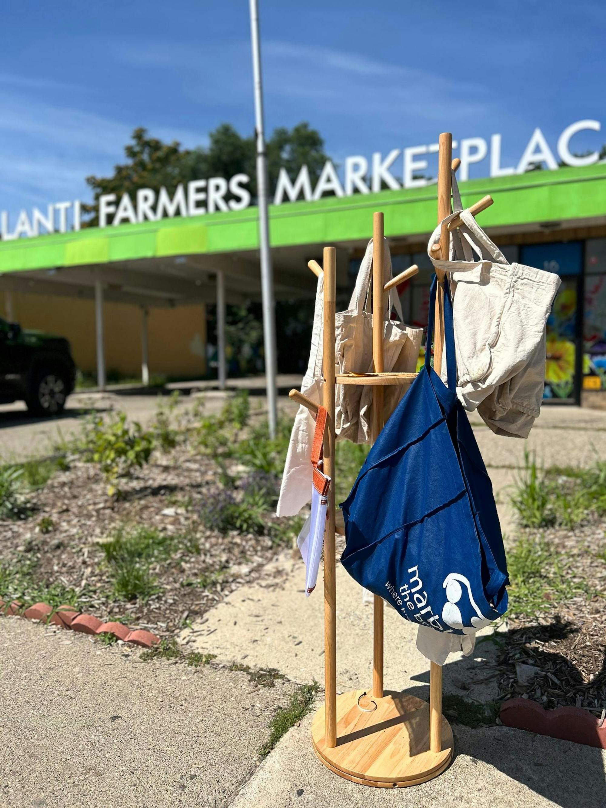 On a sunny day, several reusable cloth bags hang on a vertical wooden rack resting on the sidewalk outside of the Ypsilanti Farmers Market building.