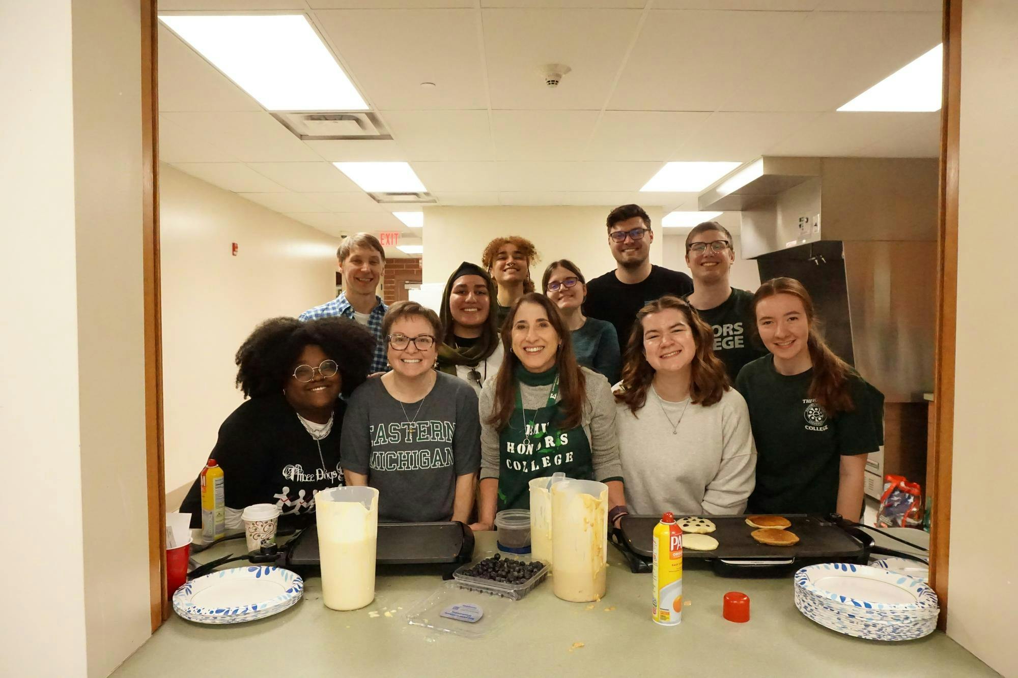 EMU students and faculty pose together behind griddles and pancake mix, ready to serve students and staff at the Honors College Pancake Breakfast.