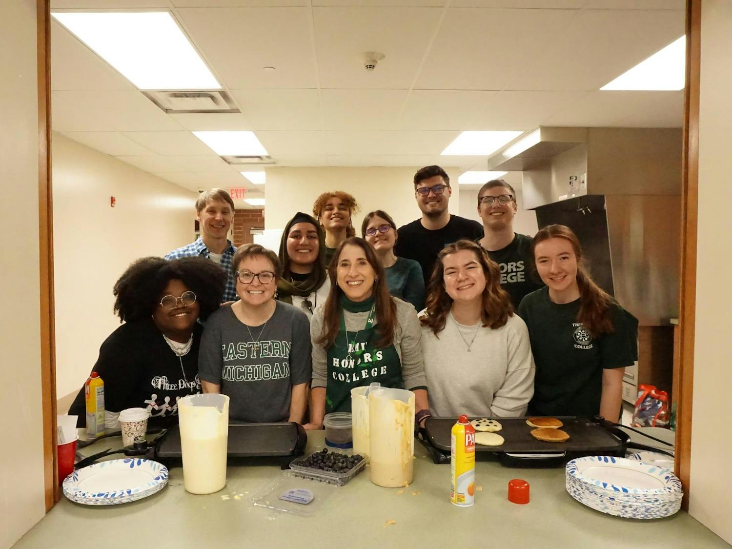 EMU students and faculty pose together behind griddles and pancake mix, ready to serve students and staff at the Honors College Pancake Breakfast.