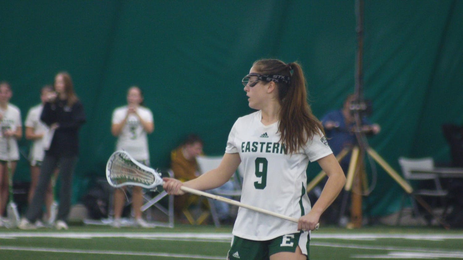 An Eastern Michigan lacrosse player in a white jersey with a green number 9 stands holding her crosse against her body while looking to the left.