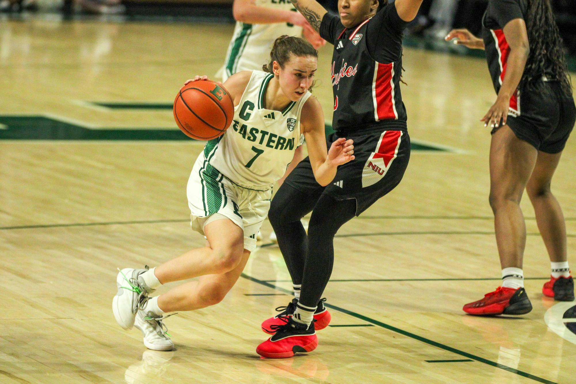 EMU player #7 Cea, in a white jersey, dribbling the basketball while an NIU player, in a black jersey, defends her. 