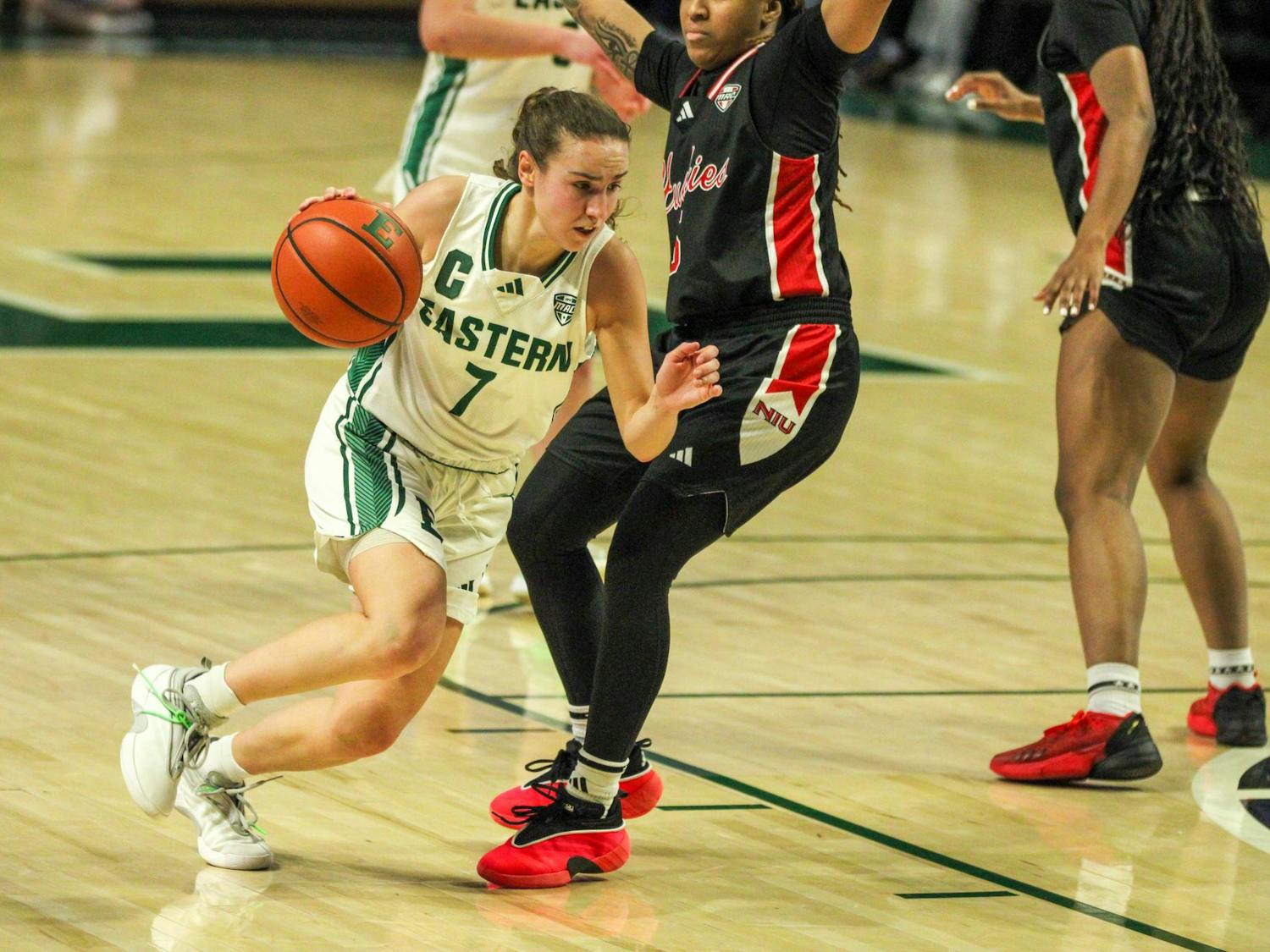 EMU player #7 Cea, in a white jersey, dribbling the basketball while an NIU player, in a black jersey, defends her.