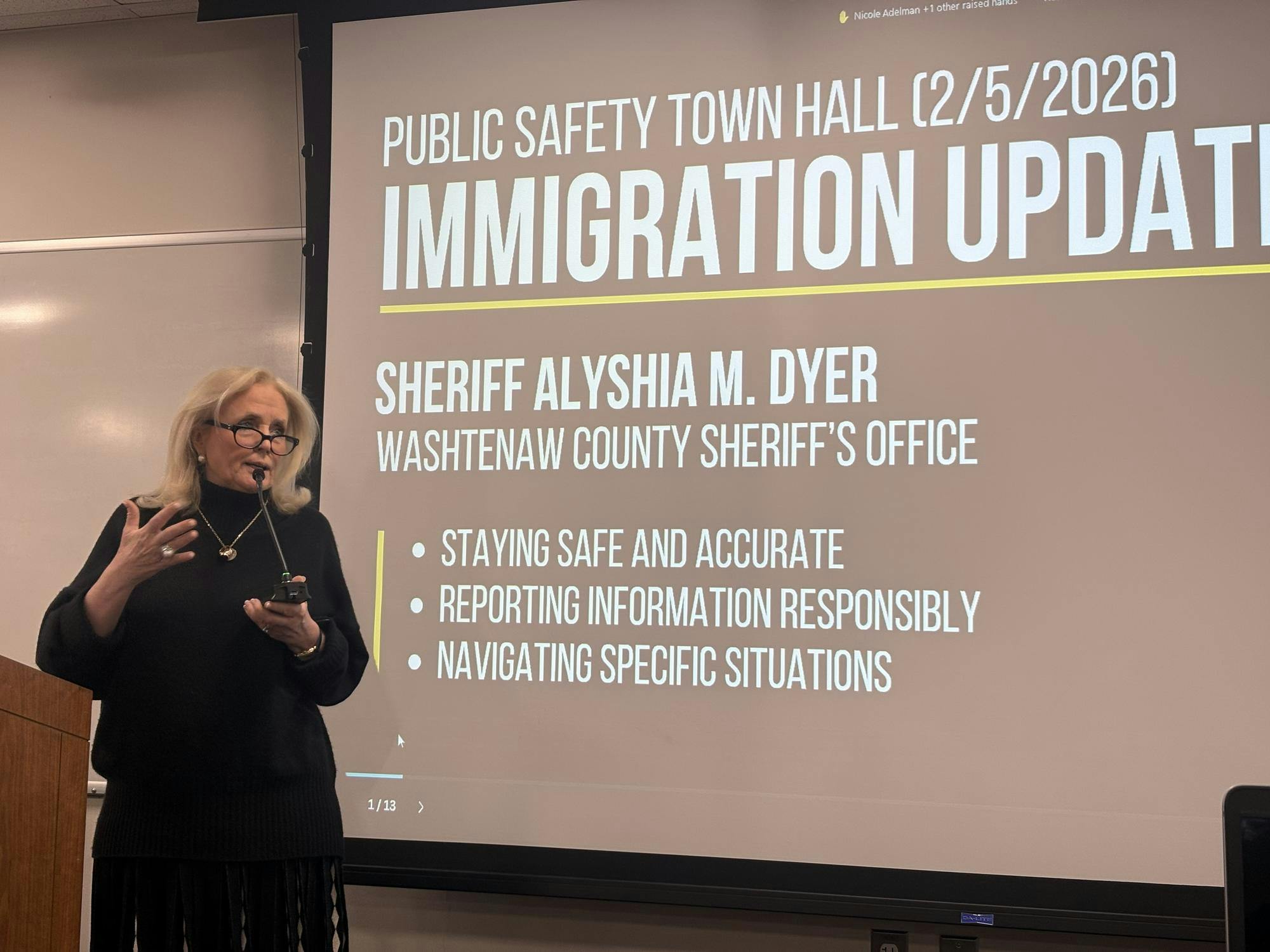 Debbie Dingell speaks into a microphone and stands in front of a projector screen that says "Public Safety Town Hall: Immigration Update."