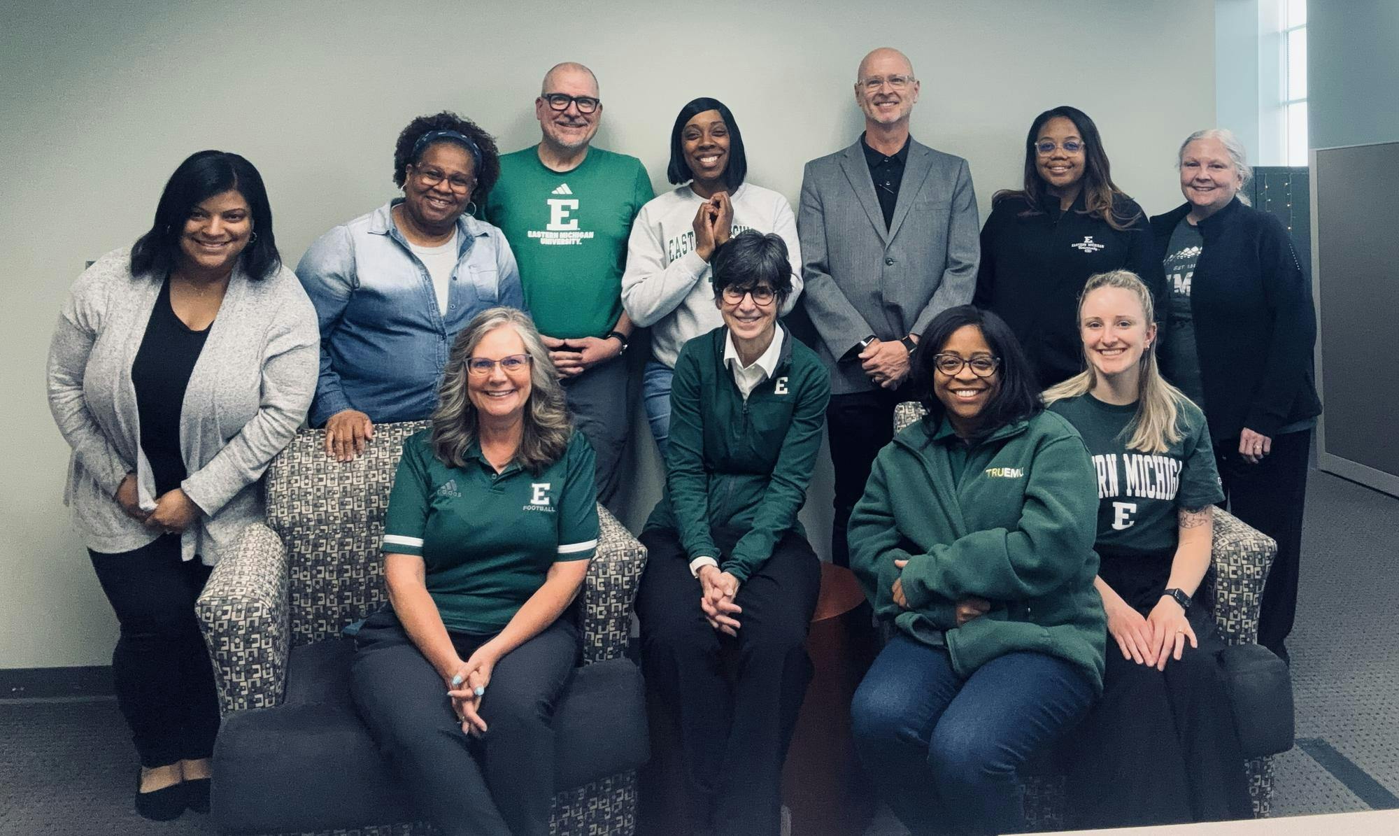 A group of people, many wearing EMU logo clothing, smile for a photo in an office space.