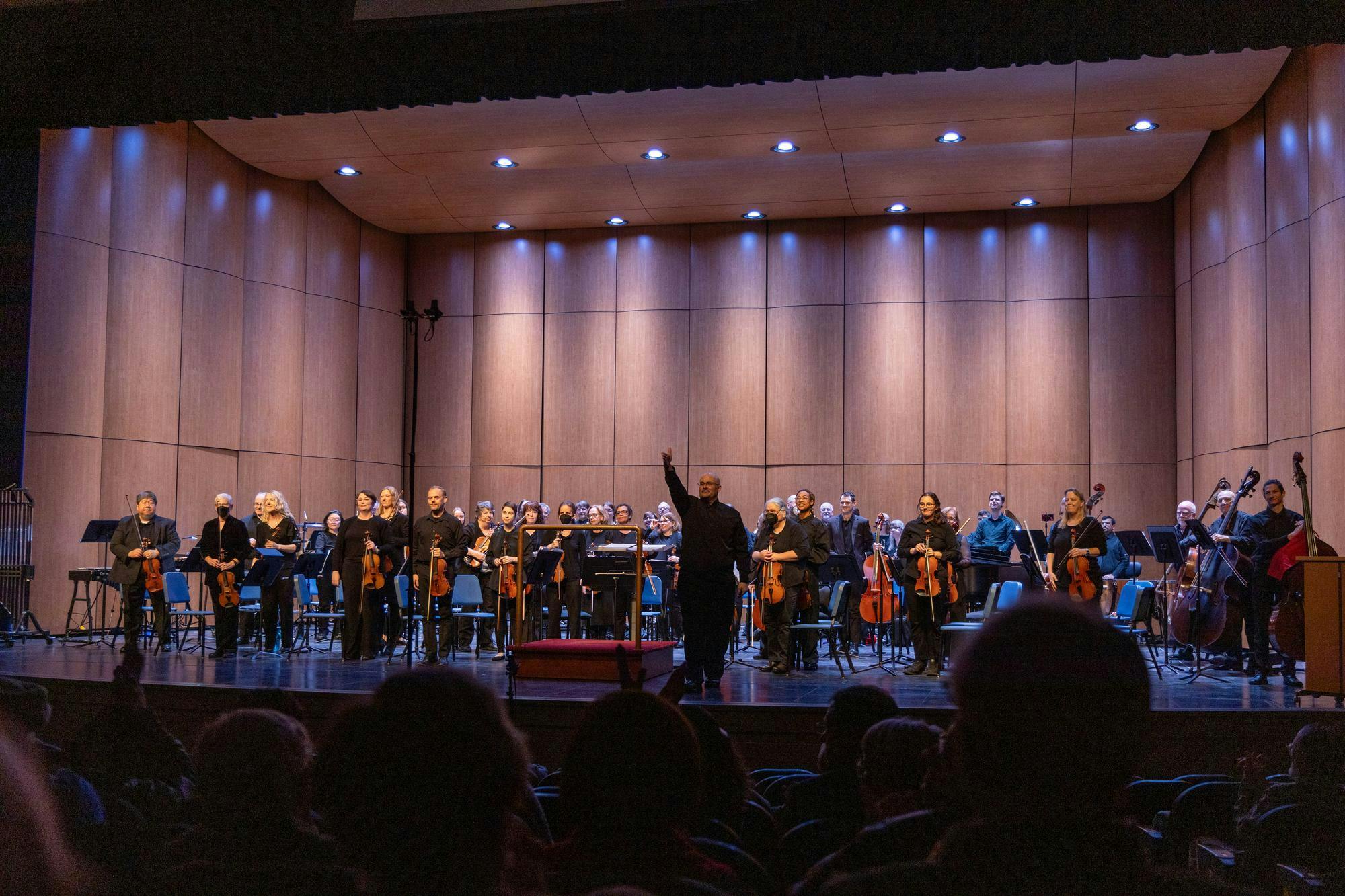 Adam Riccinto, the music director and founder of the Ypsilanti Symphony Orchestra, is wearing all black clothing and glasses as he stands on a stage facing the audience. Standing behind him and also facing the audience are the members of the Ypsilanti Symphony Orchestra, who are likewise all dressed in black. Riccinto stands to the right of a conductor's podium.