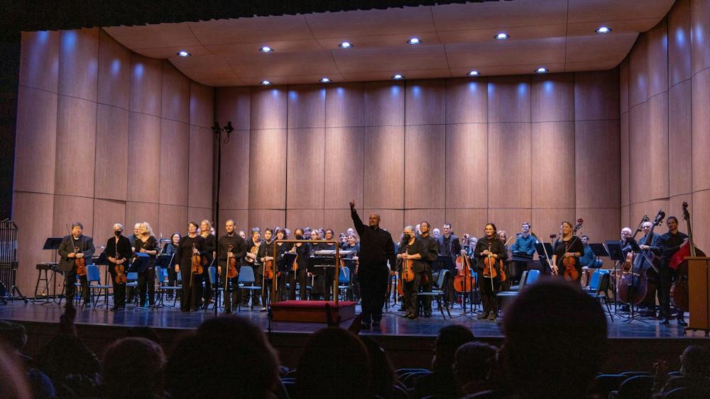 Adam Riccinto, the music director and founder of the Ypsilanti Symphony Orchestra, is wearing all black clothing and glasses as he stands on a stage facing the audience. Standing behind him and also facing the audience are the members of the Ypsilanti Symphony Orchestra, who are likewise all dressed in black. Riccinto stands to the right of a conductor's podium.