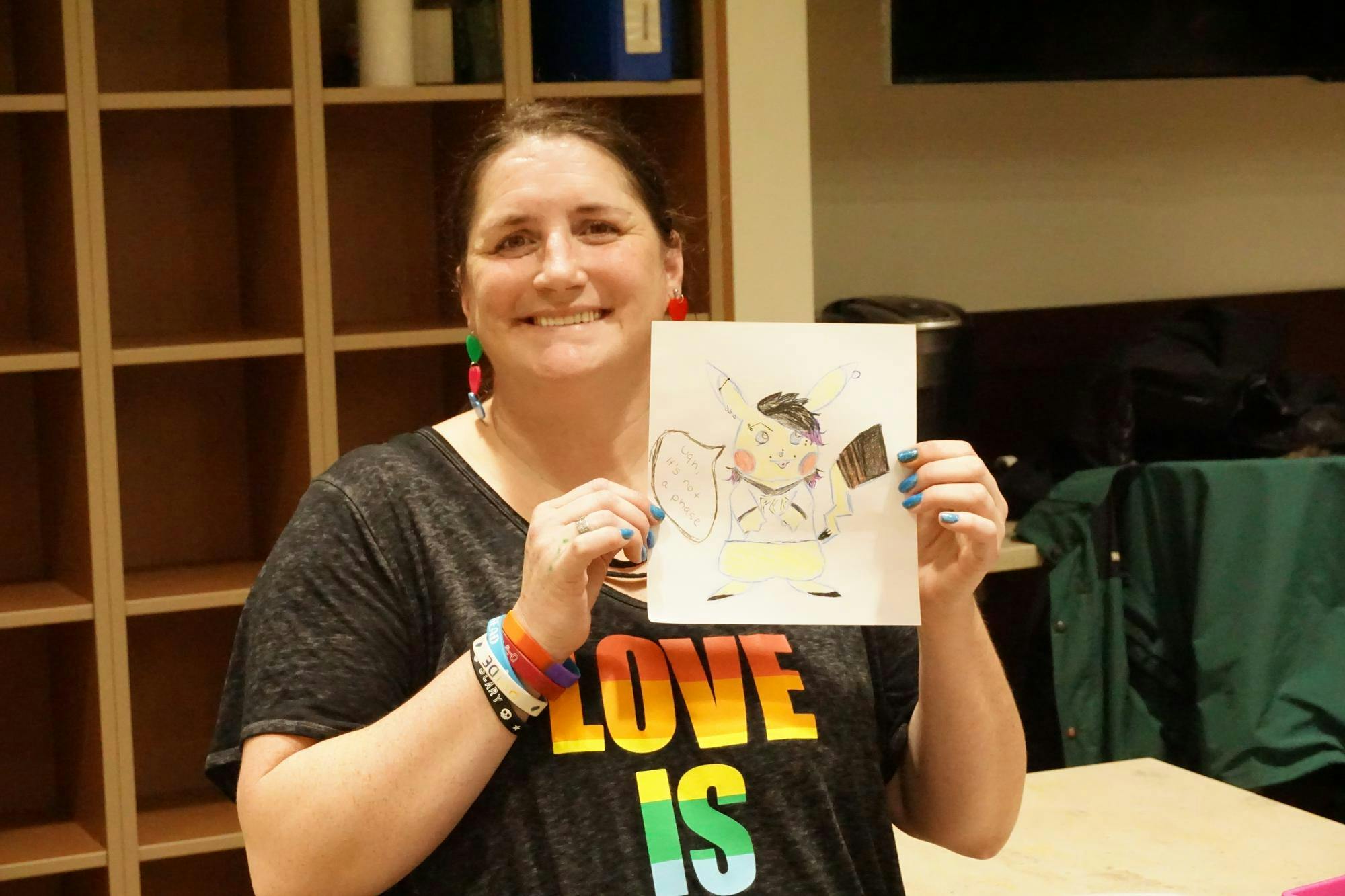 Emily Bryson is smiling while holding up a collaborative drawing featuring an emo-style Pikachu from the Found Family Feast event.
