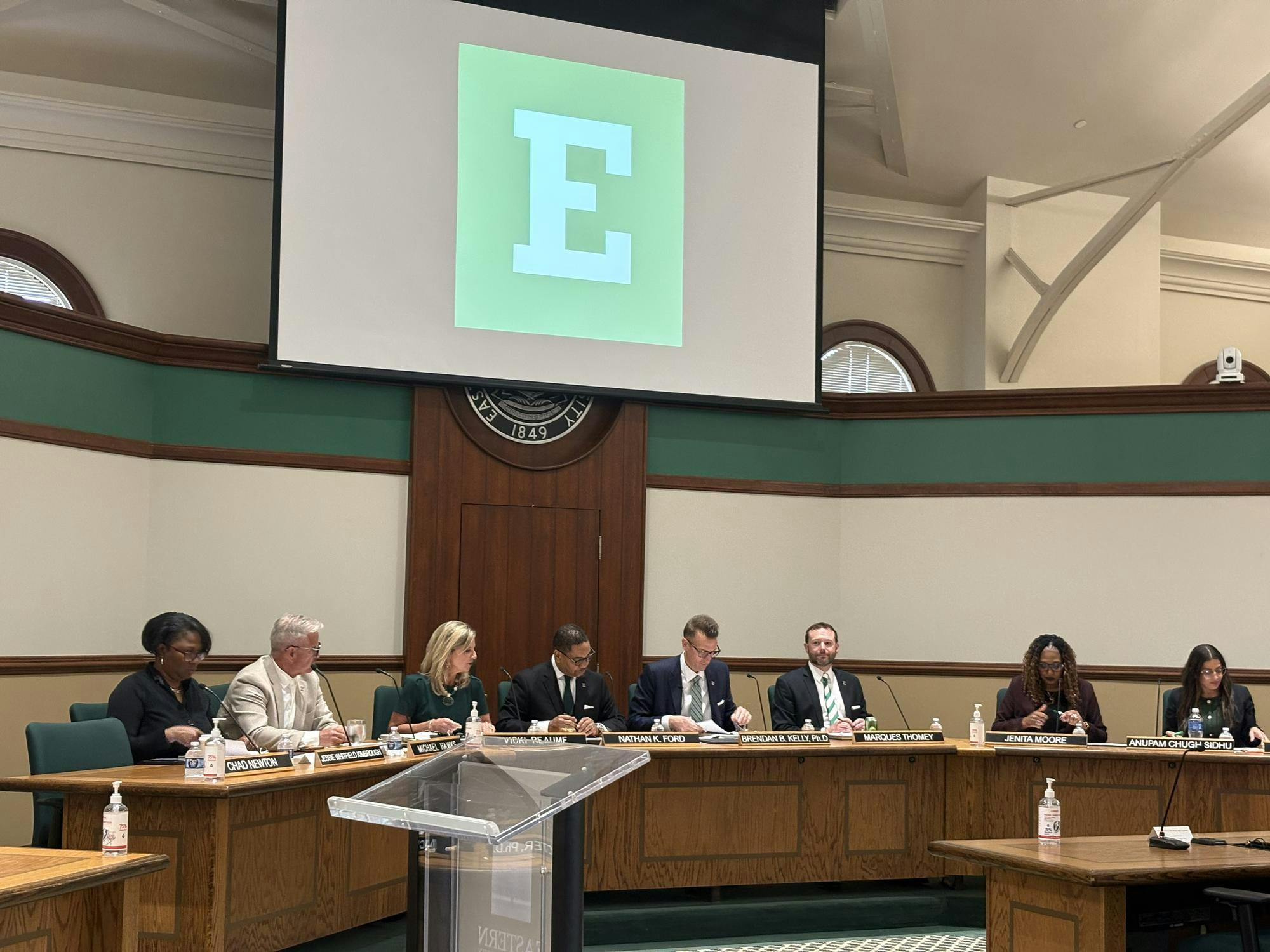 Board of Regents members sit at a long, wooden shared desk with a projector screen above them.