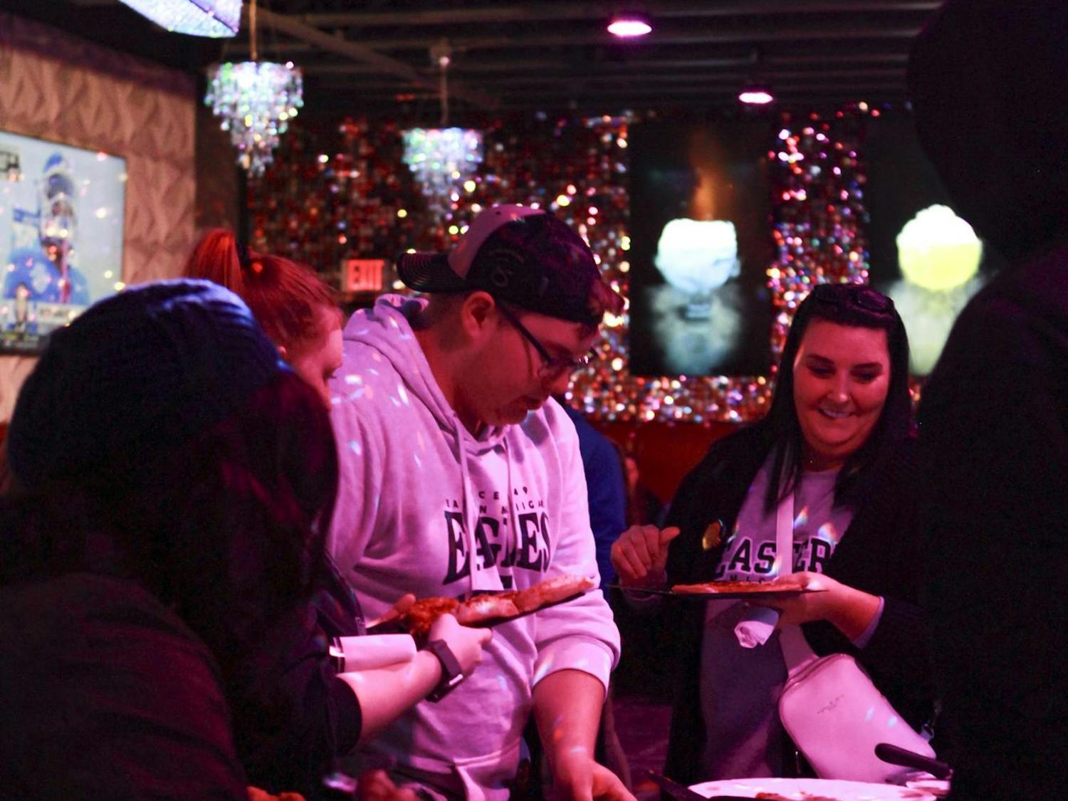 Four people are shown, two facing the camera, as they fill their plates with pizza in a dimly lit room with sparkly decorations and a small chandelier in the background. The two attendees facing the camera wear EMU shirts.