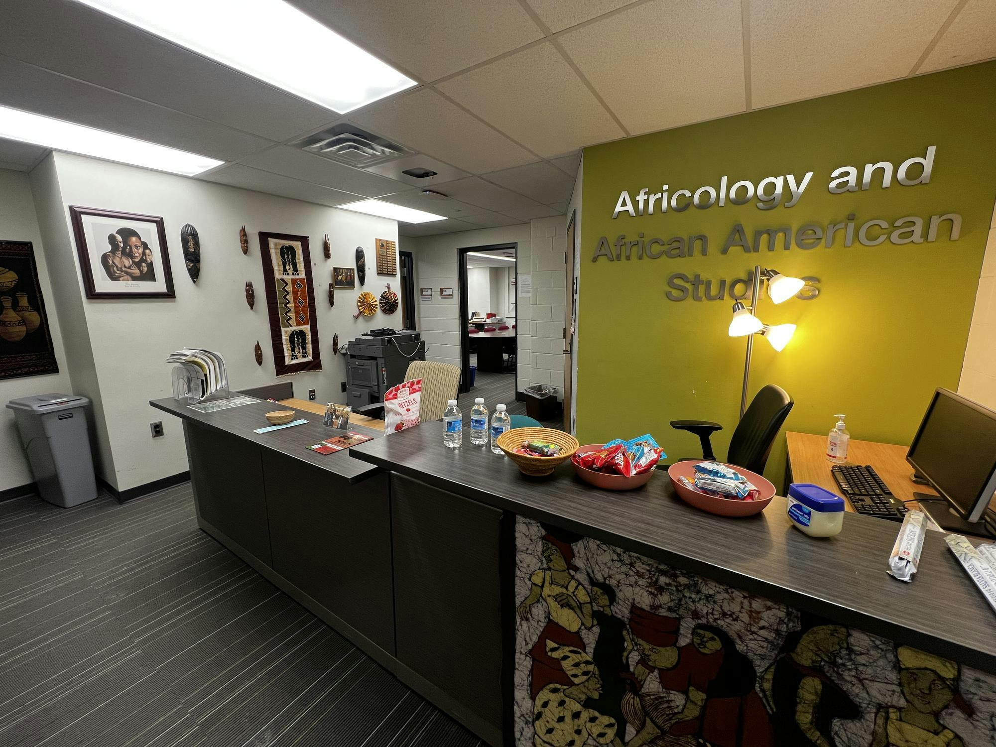 A long desk sits in front of lettering on the wall reading "Africology and African American Studies." There are various snacks on the desk. African artwork is displayed in various parts of the room.