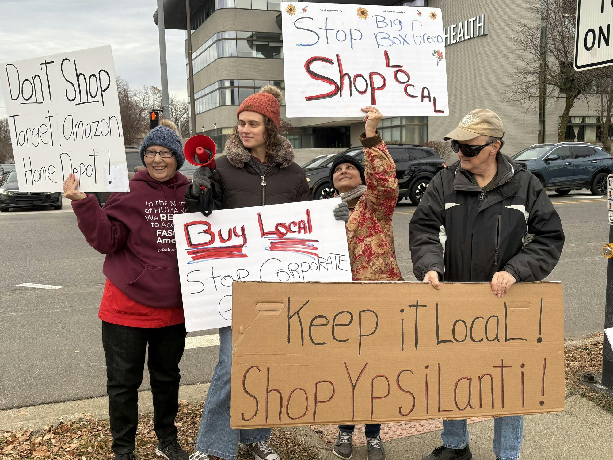 4 people stand on a sidewalk in front of a busy road. They hold cardboard signs that encourage onlookers to shop locally.