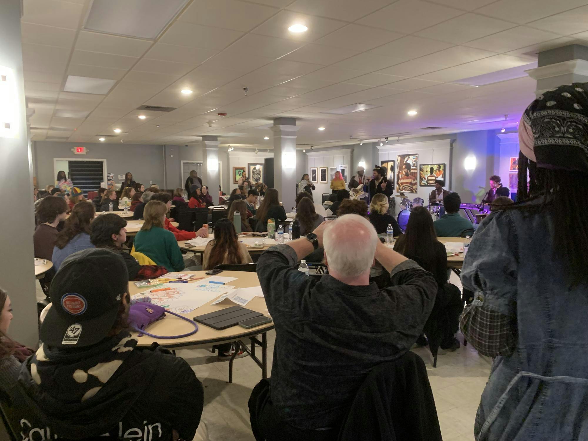Dozens of attendees sit in front of The Songbirds, a band, while they perform inside a large gallery room. 