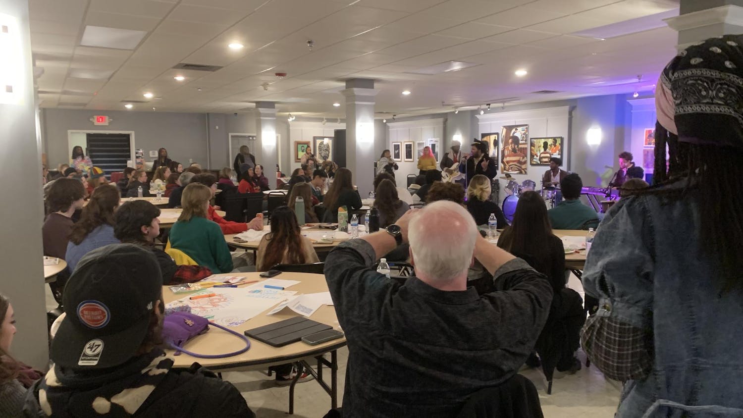 Dozens of attendees sit in front of The Songbirds, a band, while they perform inside a large gallery room.