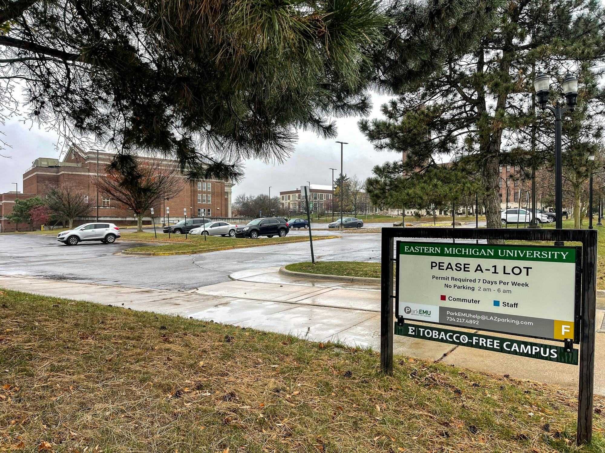 Parking lot on a rainy day. A few cars are parked, and 3 red, brick campus buildings are in the background. Parking sign reads "Pease A-1 Lot, Permit required 7 days per week."