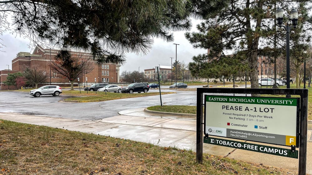 Parking lot on a rainy day. A few cars are parked, and 3 red, brick campus buildings are in the background. Parking sign reads "Pease A-1 Lot, Permit required 7 days per week."