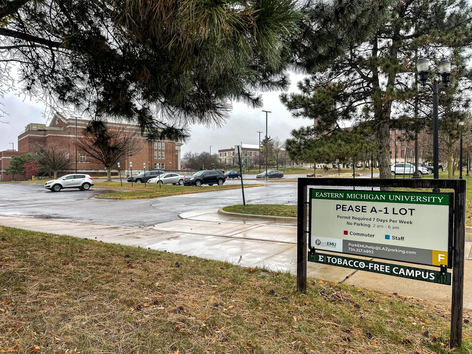 Parking lot on a rainy day. A few cars are parked, and 3 red, brick campus buildings are in the background. Parking sign reads "Pease A-1 Lot, Permit required 7 days per week."