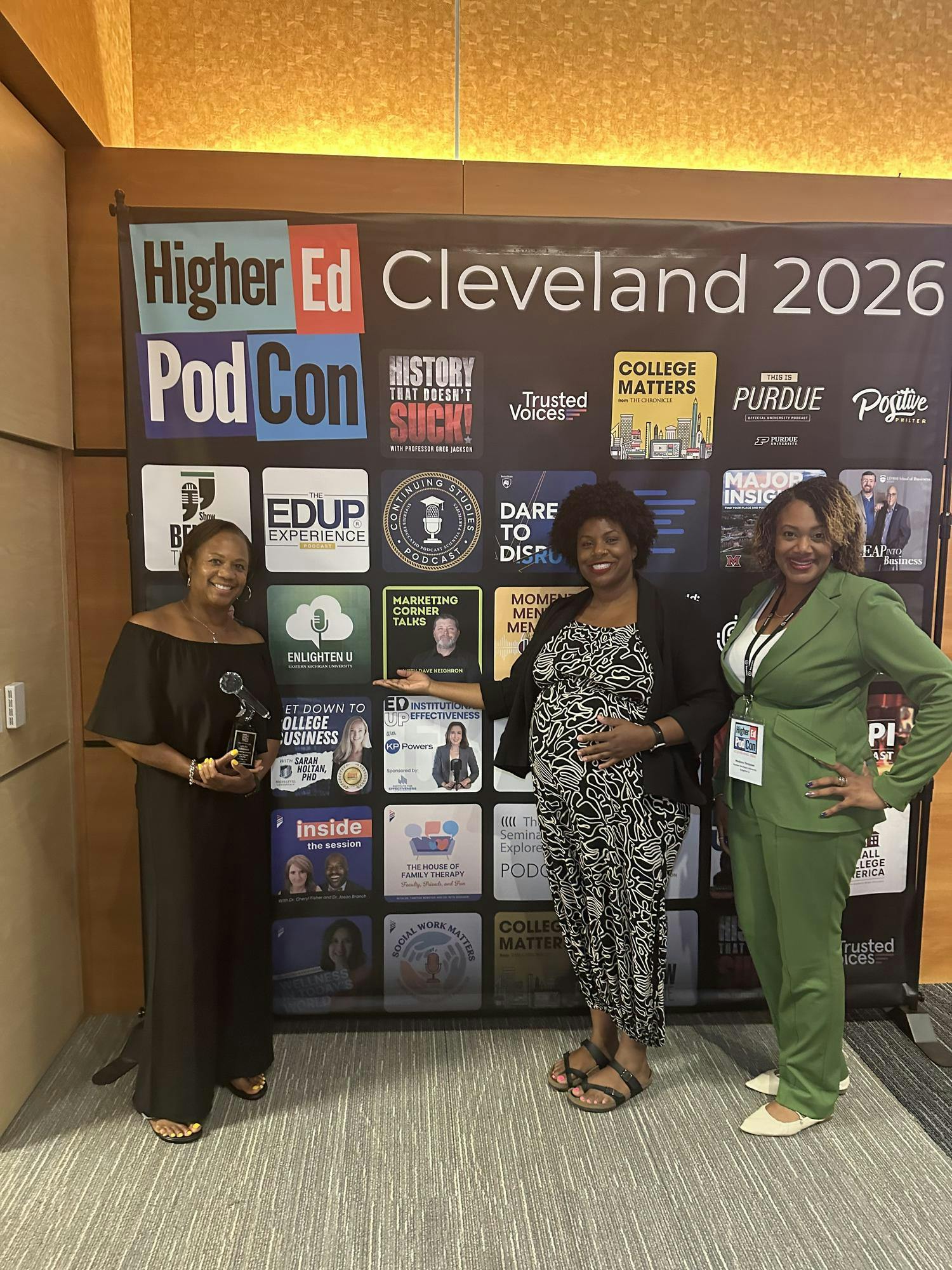 Three women standing in front of the HigherEd PodCon 2026 poster that includes the logos of various podcasts.