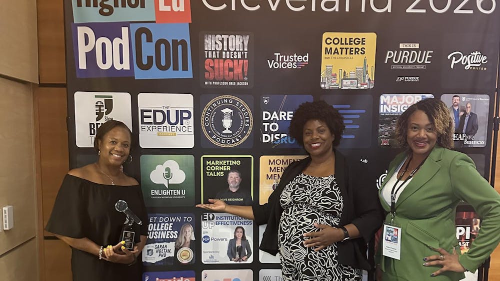 Three women standing in front of the HigherEd PodCon 2026 poster that includes the logos of various podcasts.