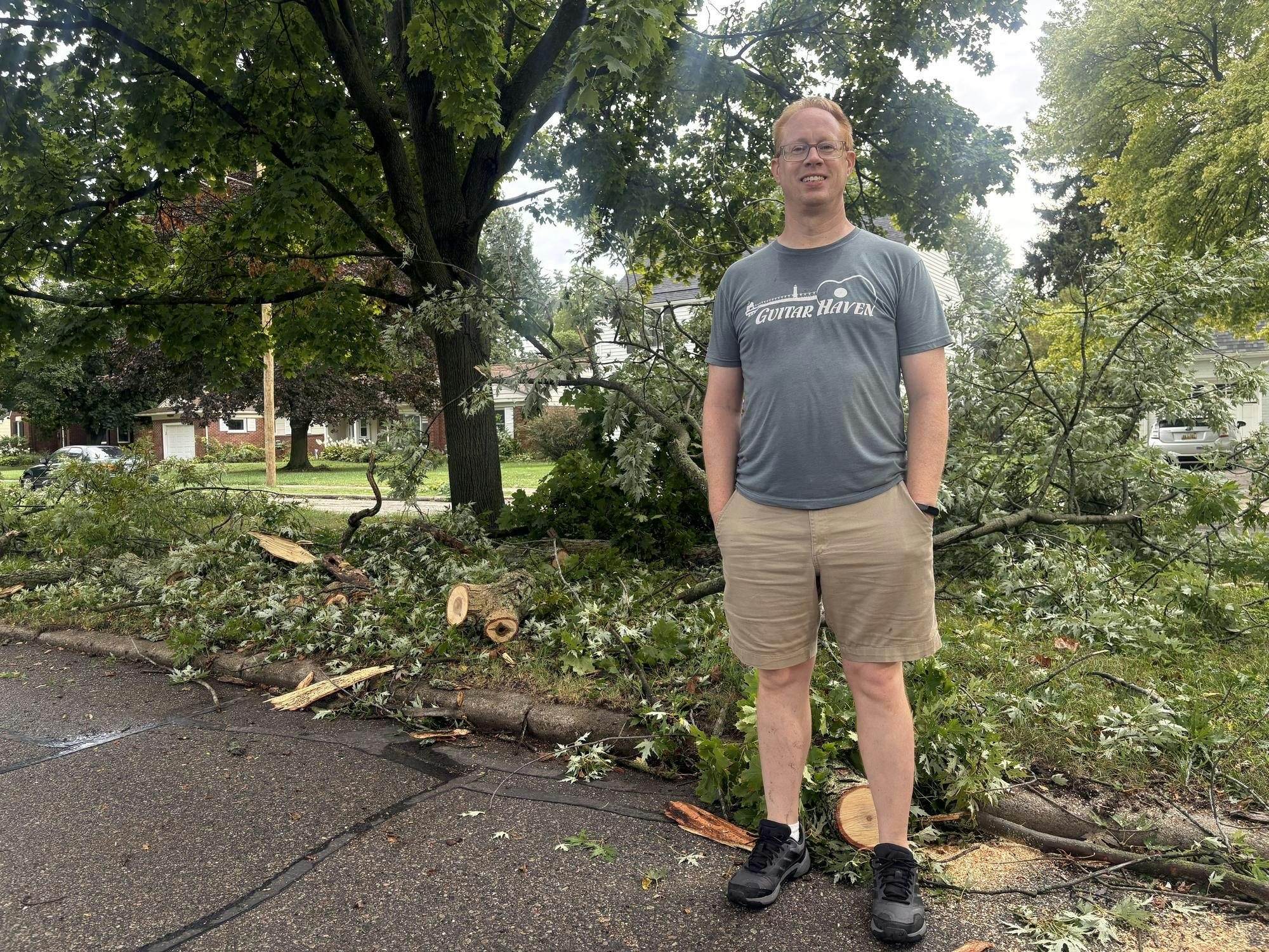 A man stands in front of several fallen tree limbs sitting on the median of a residential road.