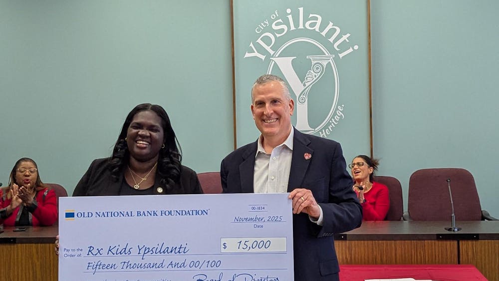 Two people hold a very large sign resembling a check for $15,000. People cheer while sitting at a long desk behind them.