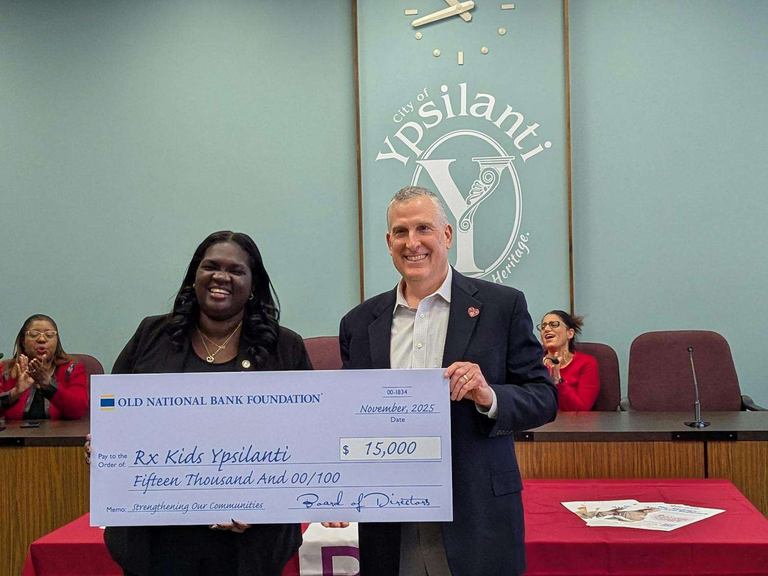 Two people hold a very large sign resembling a check for $15,000. People cheer while sitting at a long desk behind them.