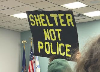 A rectangular black sign is held in the air, in bright green, bold letters is the slogan "shelter not police'. 