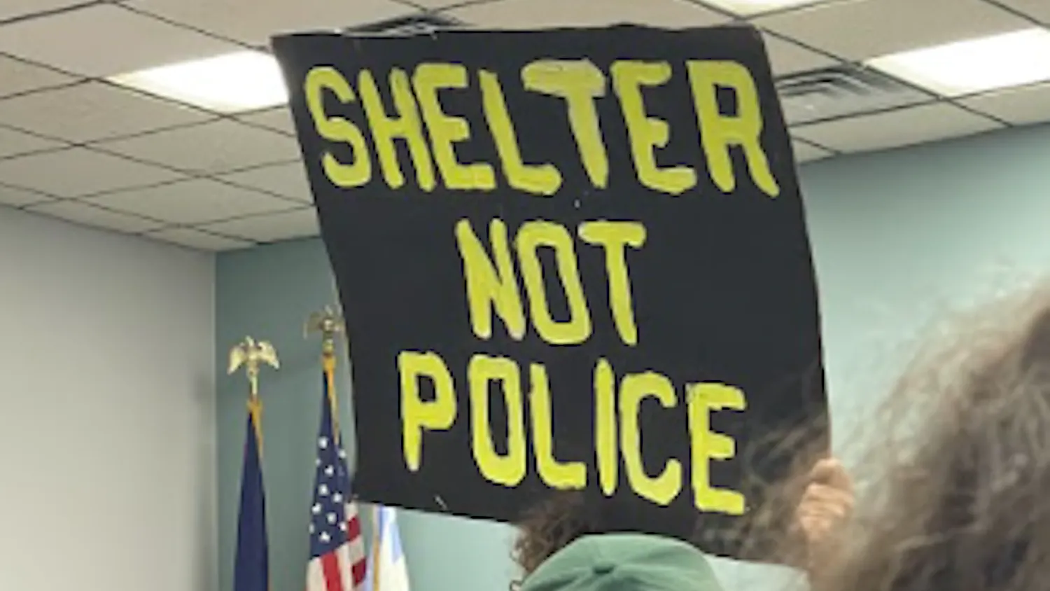 A rectangular black sign is held in the air, in bright green, bold letters is the slogan "shelter not police'.