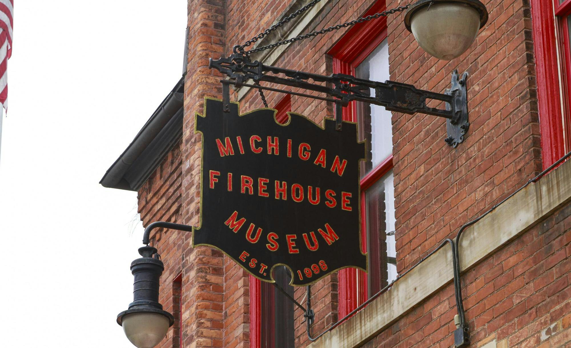 A close-up shot of a black sign with the words "Michigan Firehouse Museum Est. 1998" printed on it in red letters outlined in gold. The sign itself is also outlined in gold. The sign is sticking out from the side of a red brick building.