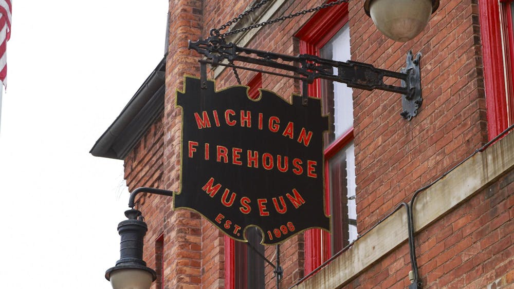 A close-up shot of a black sign with the words "Michigan Firehouse Museum Est. 1998" printed on it in red letters outlined in gold. The sign itself is also outlined in gold. The sign is sticking out from the side of a red brick building.
