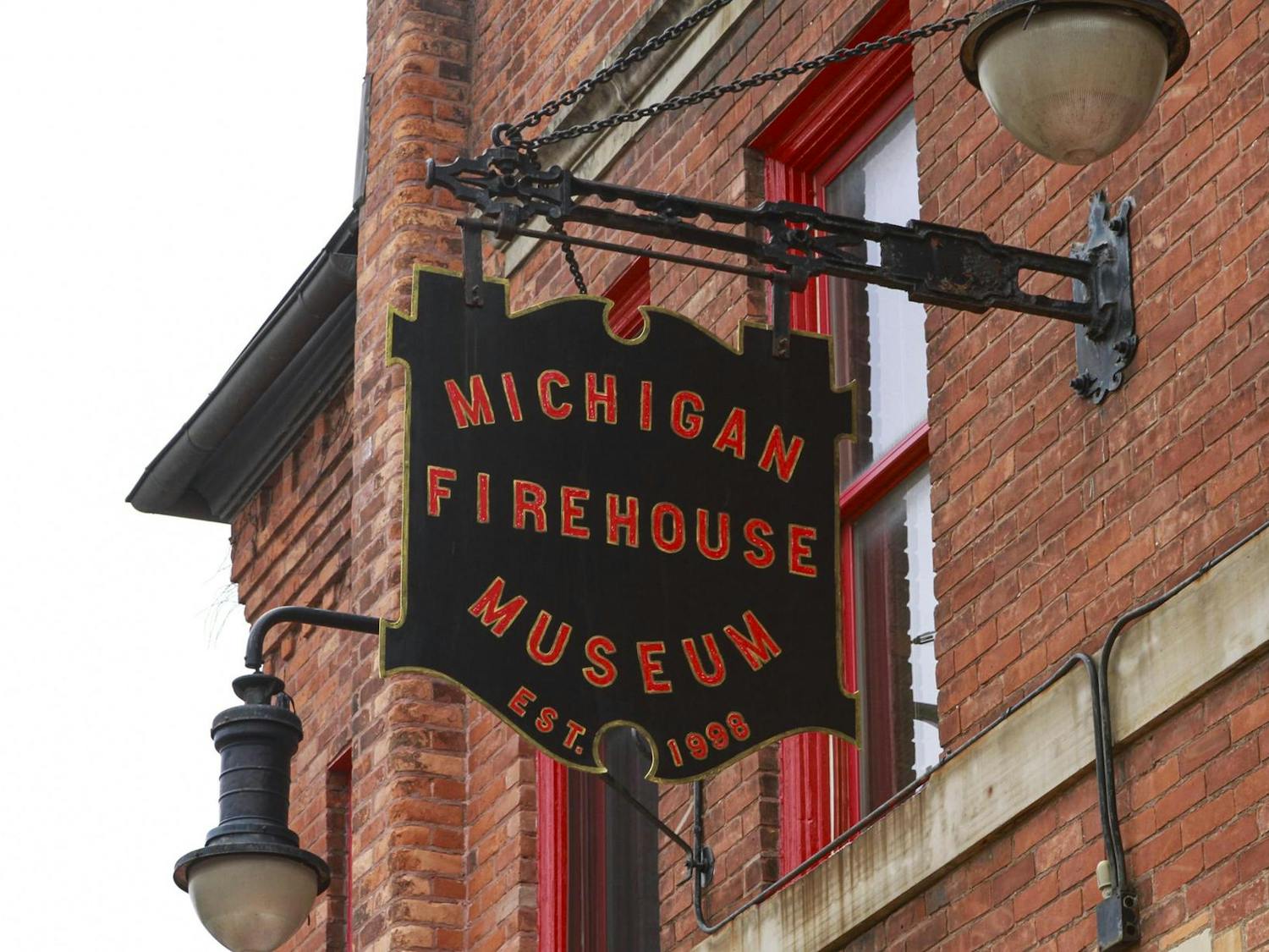 A close-up shot of a black sign with the words "Michigan Firehouse Museum Est. 1998" printed on it in red letters outlined in gold. The sign itself is also outlined in gold. The sign is sticking out from the side of a red brick building.