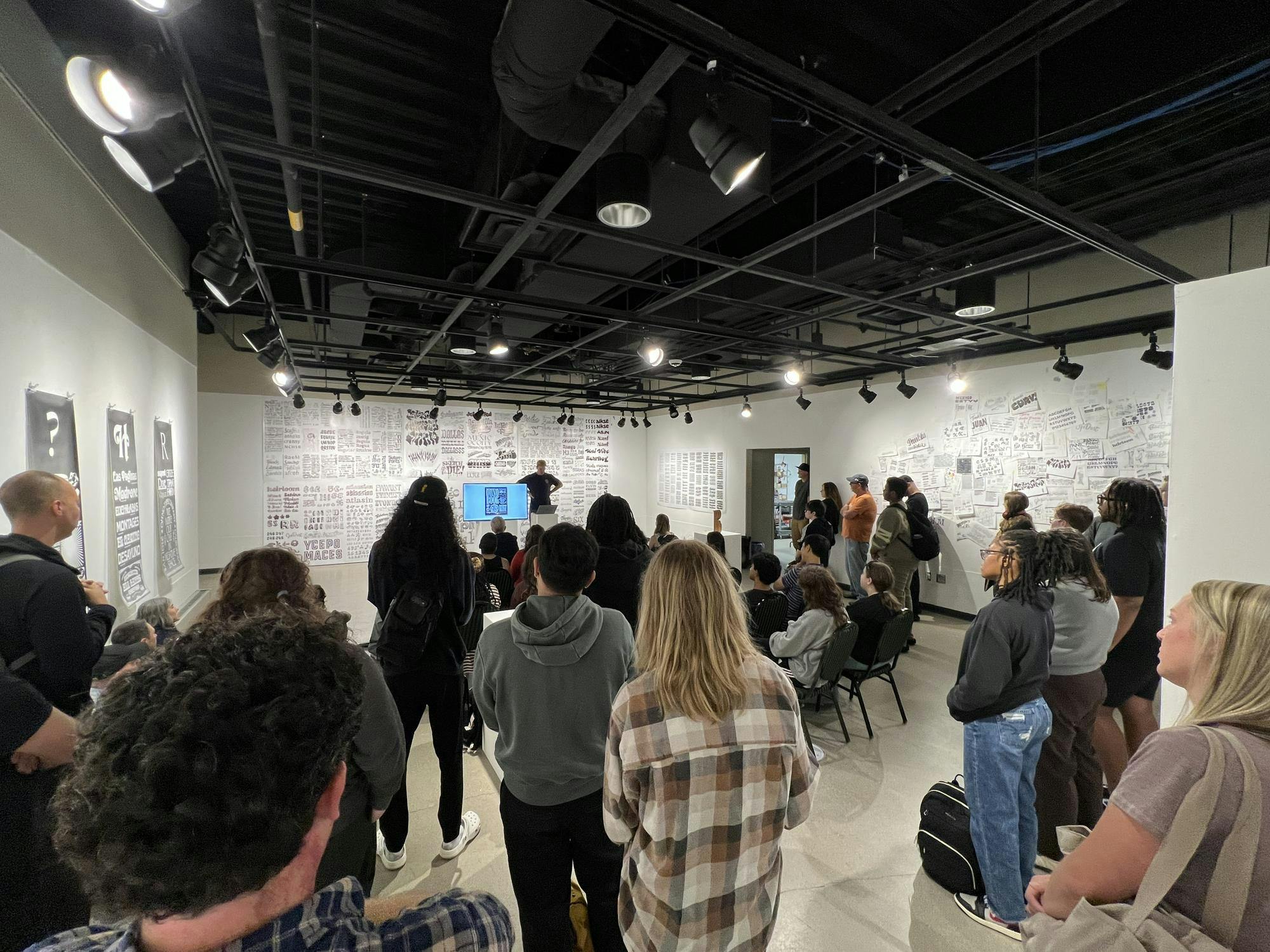 Dozens of people stand and sit wall to wall in an art gallery with papers displaying a variety of typefaces on the walls. James Edmondson is standing in the back of the room next to a TV with a presentation on it.