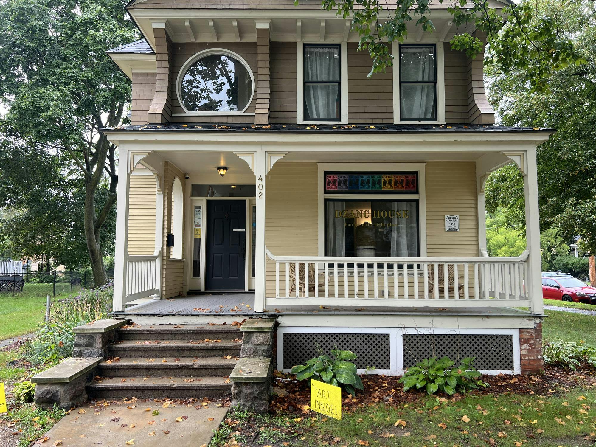 A two-story house is seen from the sidewalk, with the words "Dzanc House" on the front window.