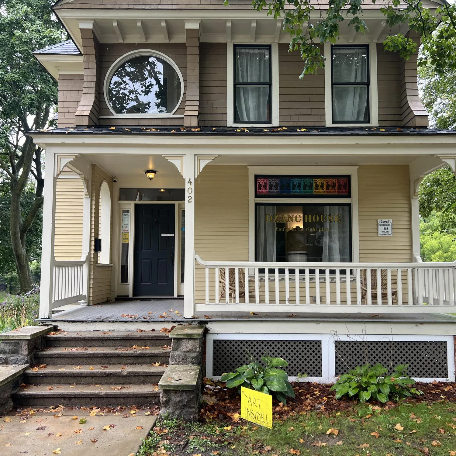 A two-story house is seen from the sidewalk, with the words "Dzanc House" on the front window.