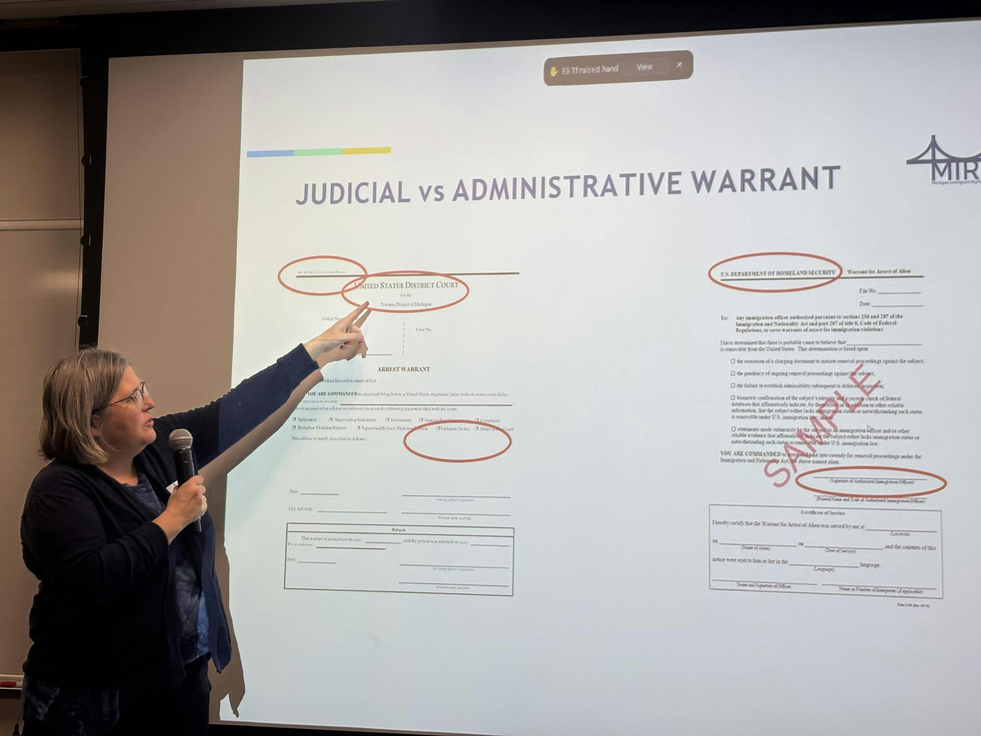 Rebecca Olszewski stands in front of a projector screen displaying a sample of a judicial warrant and an administrative warrant. Red circles on the warrants explain key differences, such as labels that say "United States District Court" on a judicial warrant and say "U.S. Department of Homeland Security" on an administrative warrant.
