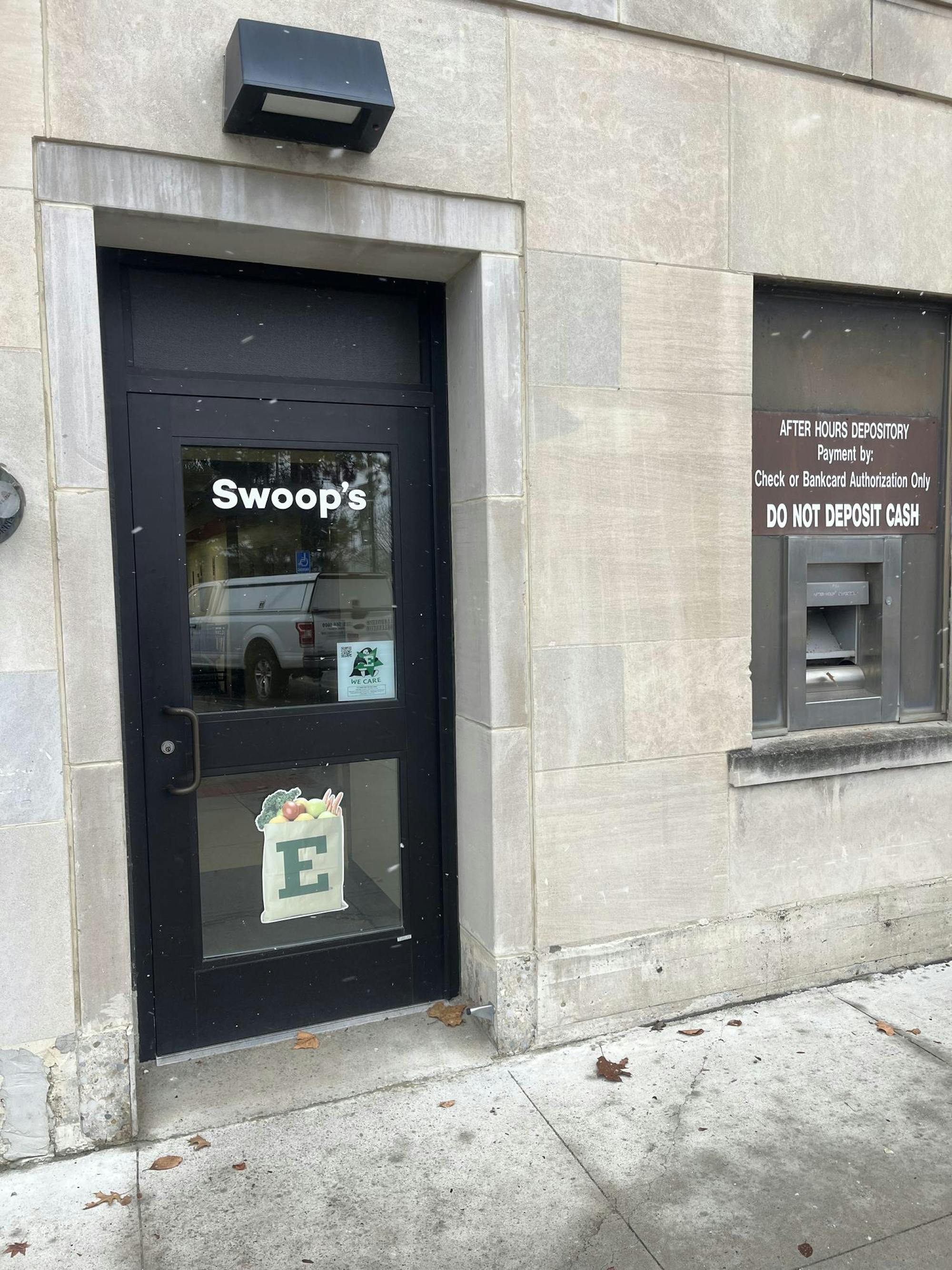 Black, windowed door reads "Swoop's" and has a sticker of an EMU-branded paper bag with groceries underneath. The door is surrounded by the gray cement of the building and an inactive cash depository.