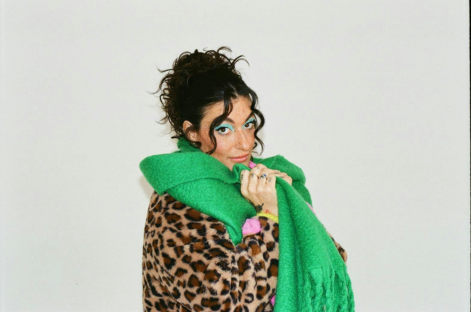Chloe Gray poses wearing a leopard print coat, a thick green scarf, and bright blue eyeshadow.