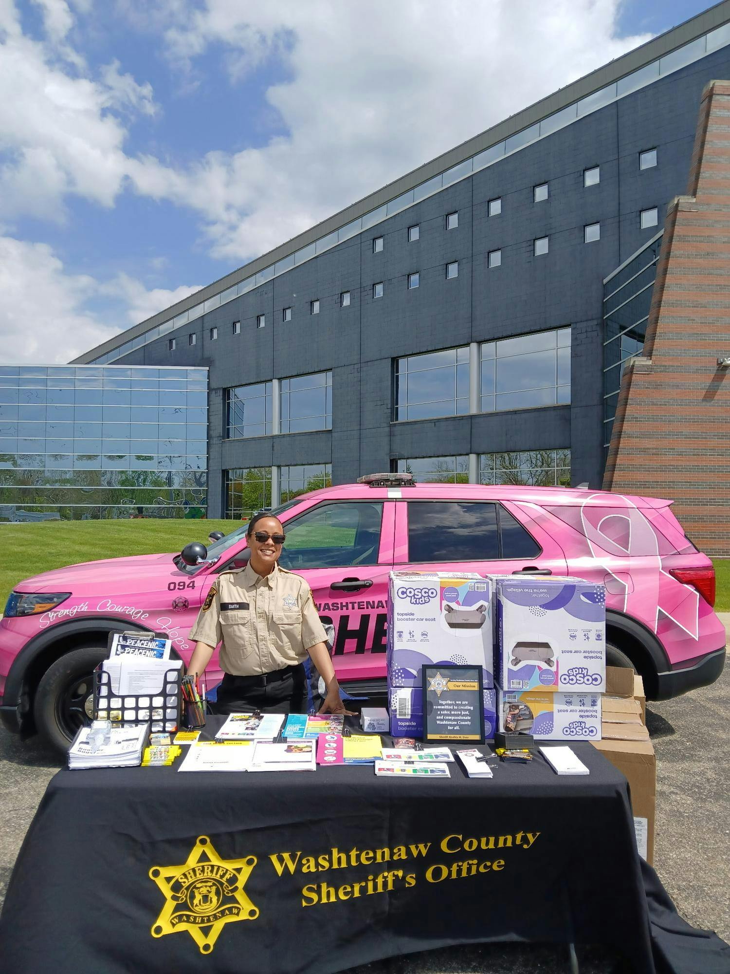 A woman stands at a table full of resources and car seats in front of a pink patrol car.