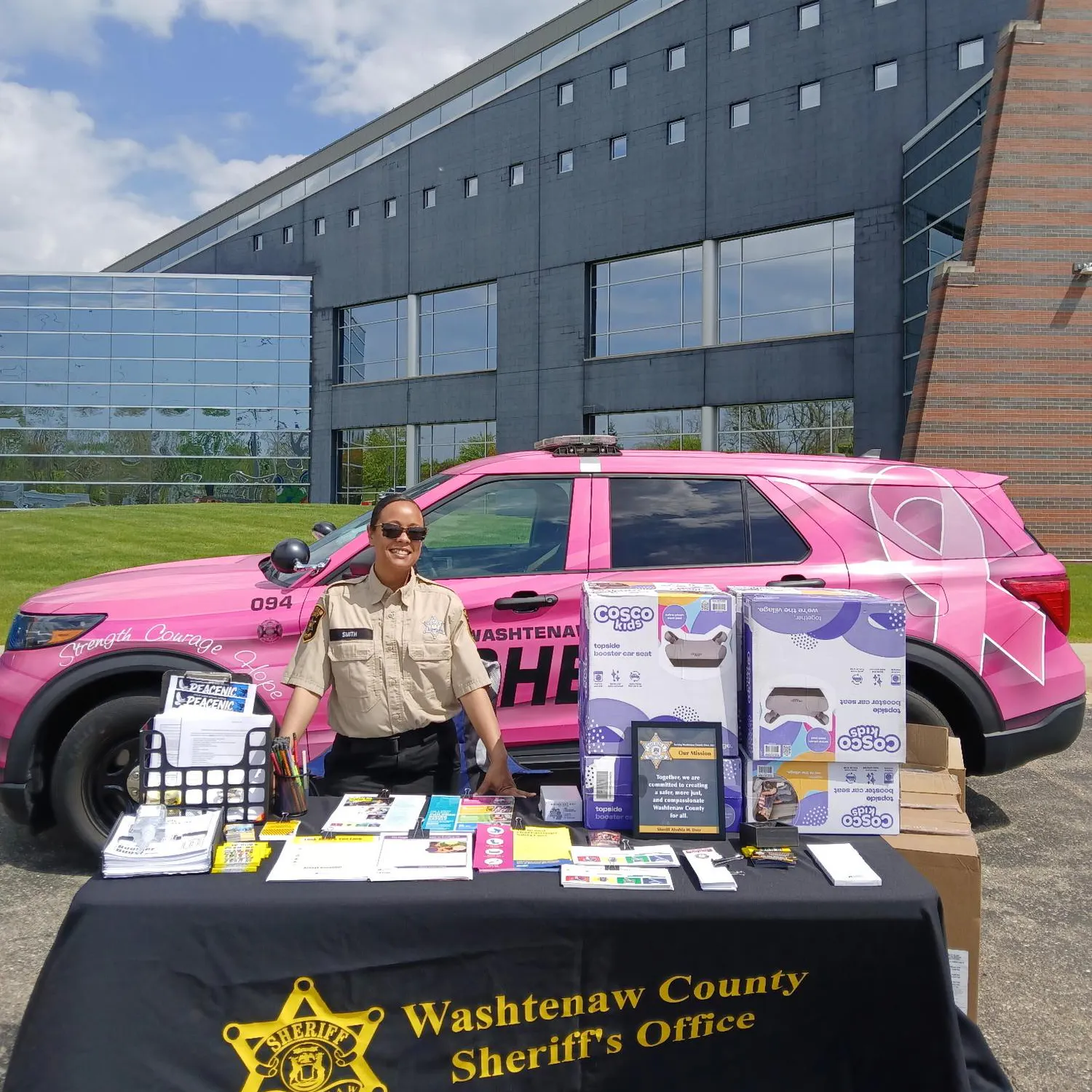 A woman stands at a table full of resources and car seats in front of a pink patrol car.