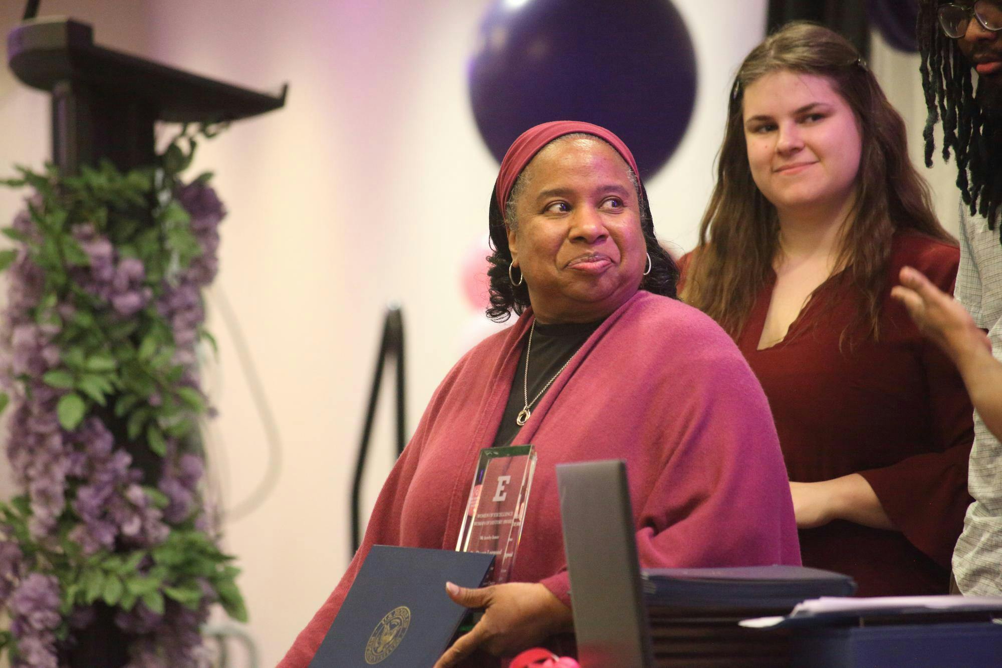 At an indoor award ceremony, a woman is standing and smiling while holding two awards. Two other people stand behind her smiling.