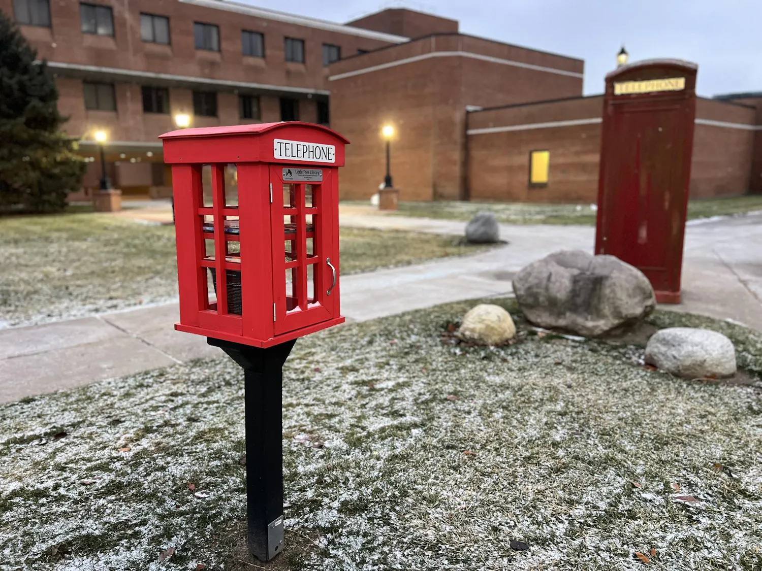 A small red box made of wood and glass stands outside on a pedestal on a snowy day. The Alexander Music Building and a red phone booth can be seen in the background. The box, which resembles the phone booth, serves as a little library and contains several books.