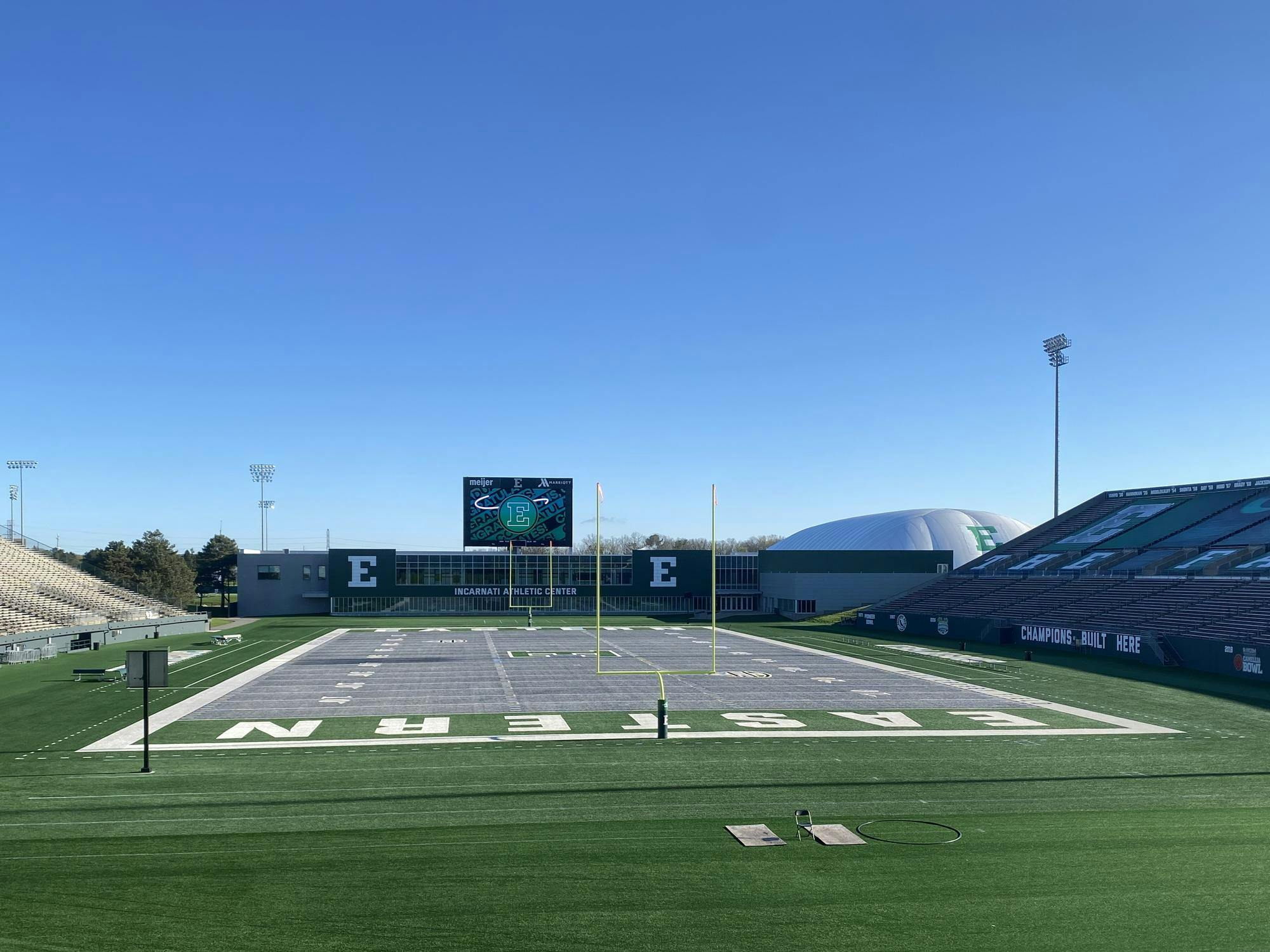 Football field with gray turf reads "Eastern" on either end zone. Empty bleachers and goalposts surround the field.