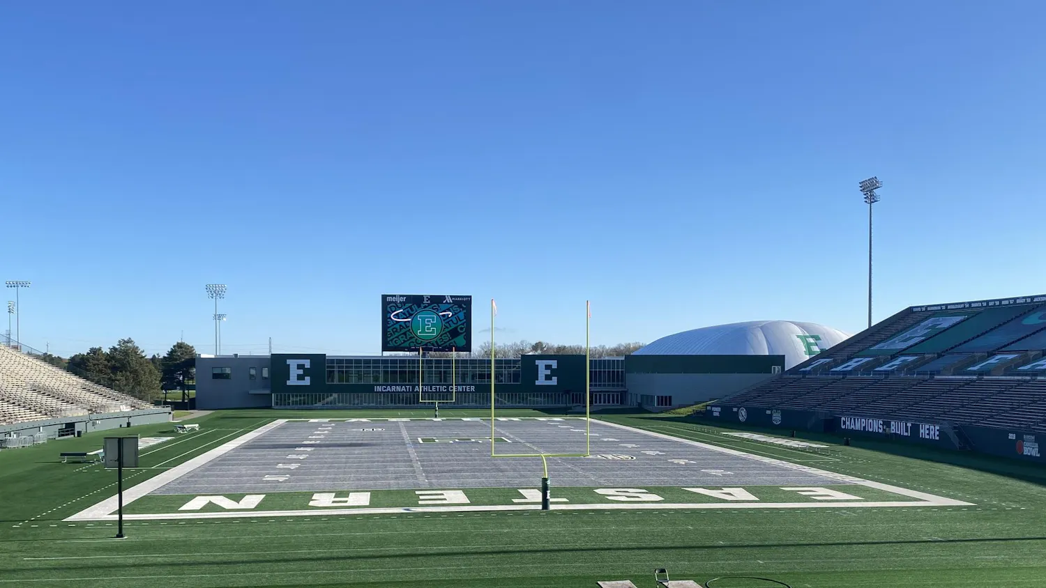 Football field with gray turf reads "Eastern" on either end zone. Empty bleachers and goalposts surround the field.