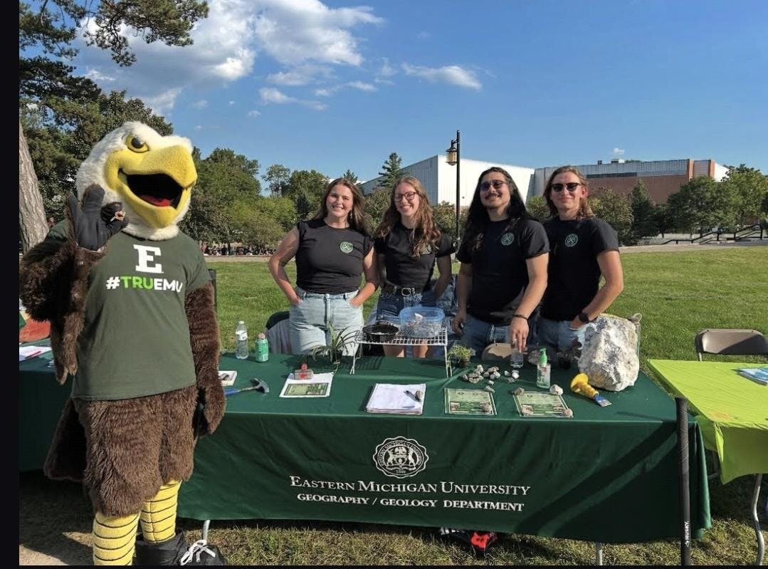 Swoop stands next to GeoClub members at their booth. There are rocks on the table. 
