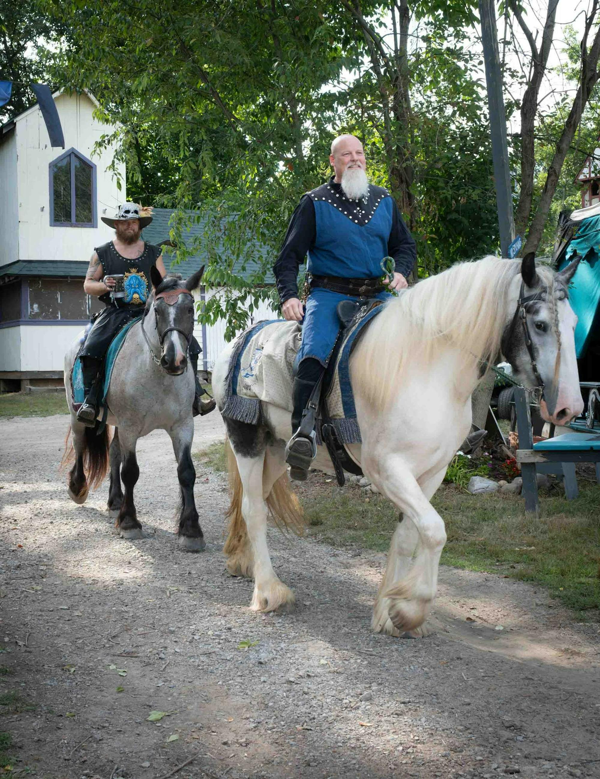 Two people dressed in renaissance attire ride horseback through festival grounds.