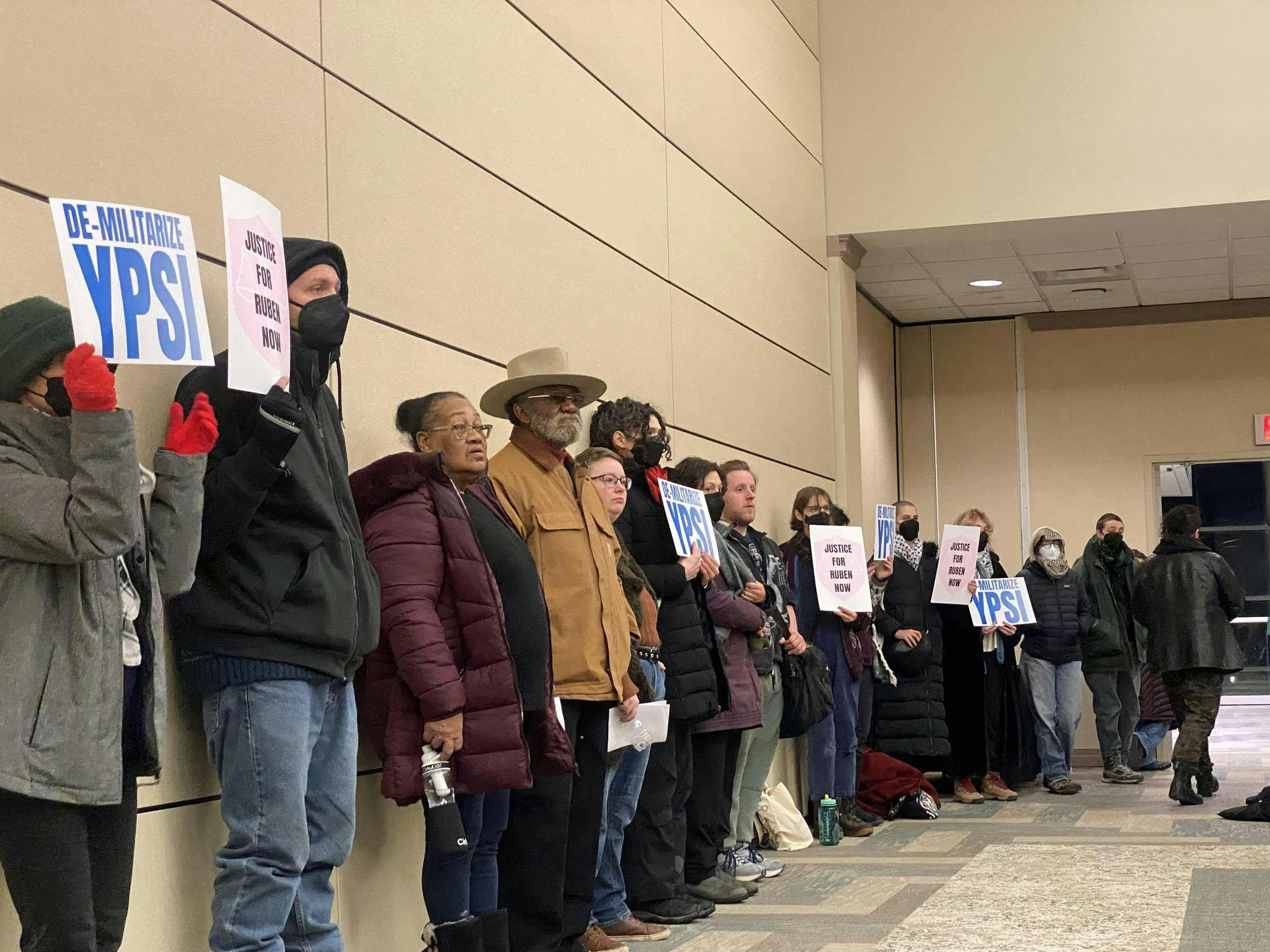 People stand against a beige wall holding signs of protest. The signs read "justice for Ruben now" and "de-militarize Ypsi."