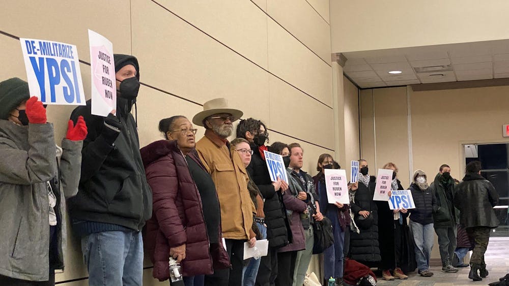 People stand against a beige wall holding signs of protest. The signs read "justice for Ruben now" and "de-militarize Ypsi."